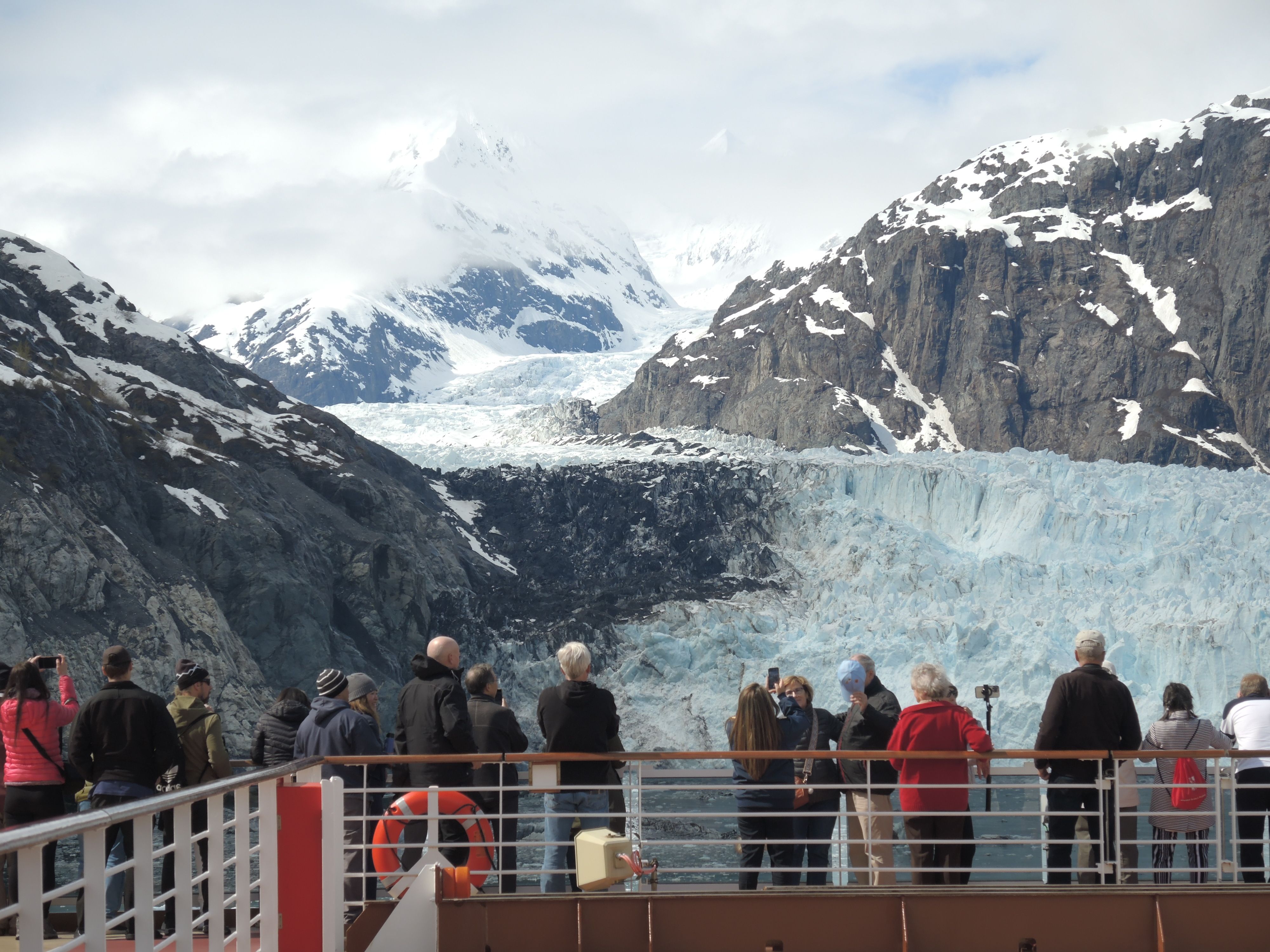 Tourists watching a glacier from a cruise ship deck surrounded by snowy mountains on an Alaska cruise